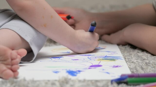 Mom helps her daughter learn to draw on paper by coloring with multi-colored pencils and felt-tip pens. Happy family playing together at home on floor. Mother, nanny, teaches child girl to draw.