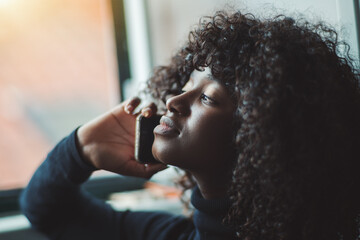 Close-up portrait with a shallow depth of field and a selective focus of a ravishing young black female with curly afro hair, talking on the phone while sitting next to the window and looking outside - obrazy, fototapety, plakaty
