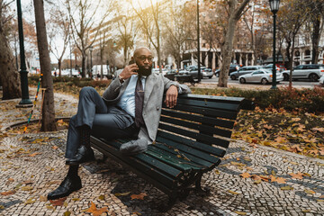 Fototapeta premium A gorgeous mature dapper bald bearded African businessman in a neat formal suit and eyeglasses is sitting on the wooden bench on the boulevard and speaking on the phone to his business partner
