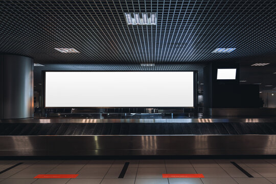 A baggage claim area in a hall of a contemporary airport arrival zone with luggage conveyor belt and a mock-up of a long white empty advertising or information billboard and tv plasma screen