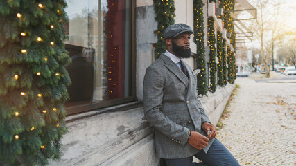 A portrait of an elegant black guy outdoors in a cap and a fashionable custom costume, with a beard, leaning against the building wall decorated with artificial Christmas tree's needles and garlands