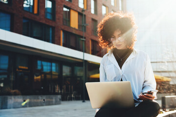 Curly haired woman is working on a laptop sitting on a bench outside in the city