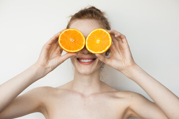 Ginger woman with freckles is covering her eyes with sliced orange and smile on a white studio wall with bare shoulders