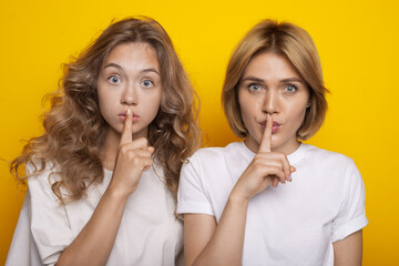Naklejka premium Two adorable women with blonde hair are gesturing at camera the silence sound posing on a yellow studio wall