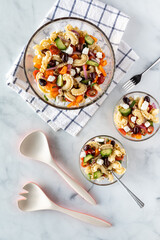 Top down view of a bowl of Greek pasta salad with two servings of the salad and salad serving utensils.