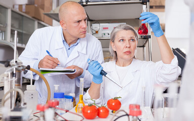 Two experienced biochemists checking fruits and vegetables for nitrates and pesticides in modern laboratory, recording experimental procedure and results..