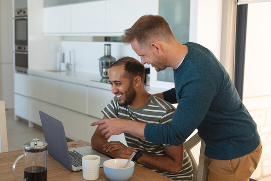 Multi ethnic gay male couple smiling and eating breakfast and using laptop at home