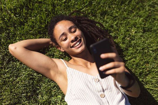 Smiling african american woman lying on grass and using a smartphone in park - Powered by Adobe