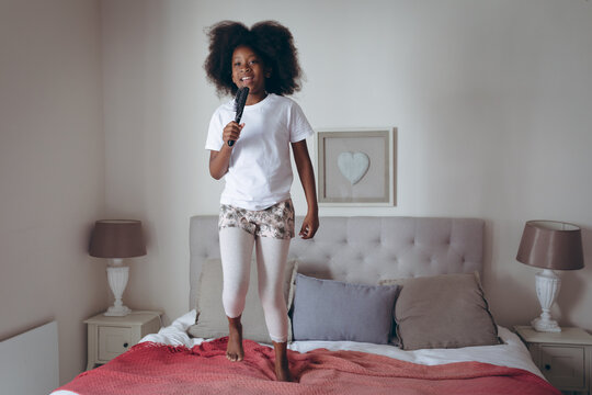 African american girl standing on bed holding hairbrush pretending to sing