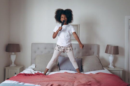 African american girl standing on bed holding hairbrush pretending to sing