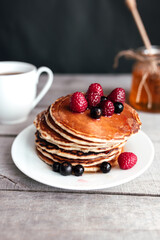 Pancakes with berries and honey on a white plate, wooden spoon, jar, coffee cup