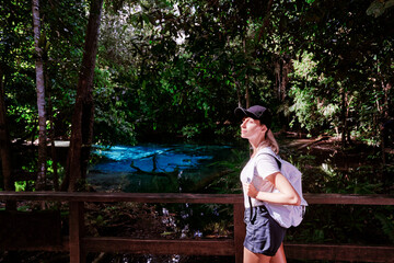 Young woman in hat with rucksack enjoying view of amazing crystal clear emerald pool in mangrove forest, Krabi, Thailand.