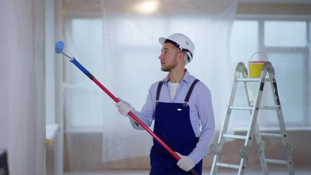 young male house painter in work suit and safety helmet makes repairs indoors and paints walls using construction roller background of window and stairs