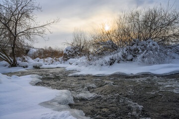Winter landscape with a stormy river on a sunny frosty evening. Beautiful branched trees on the banks, some of them covered with white frost. Winter sun and yellow colors in the sky 