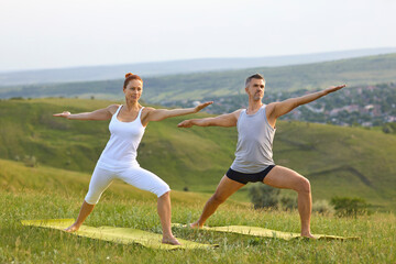Middle aged couple doing Warrior II pose while practicing yoga on grassy hill in countryside