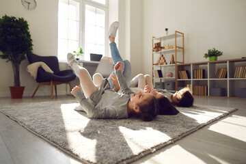 Spending time at home with children, happy family concept. Happy smiling mother and two small daughters lying on floor with legs raised up and having fun together at home in sunlight