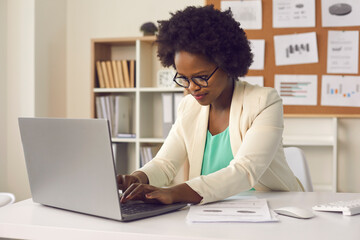 Attractive busy young african american woman sitting at desk in office, working with project report...