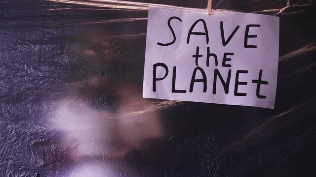 Save the planet. Climate change. Global warming. Defocused activist woman silhouette with green plant behind creased texture polyethylene film with environmental inscription banner on dark background.