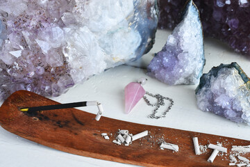 A close up image of burning incense stick with several amethyst crystals on a white table top. 