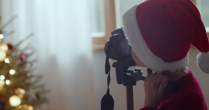 Young kid in santa claus hat shooting Christmas tree on professional camera at home background. Back view of faceless boy making photo video indoors slow motion. Festive memories equipment technology