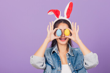 Happy beautiful young woman smiling wearing rabbit ears and denims holding colorful Easter eggs front eyes, Thai female with bunny ear, easter egg cover eye, studio shot isolated on purple background