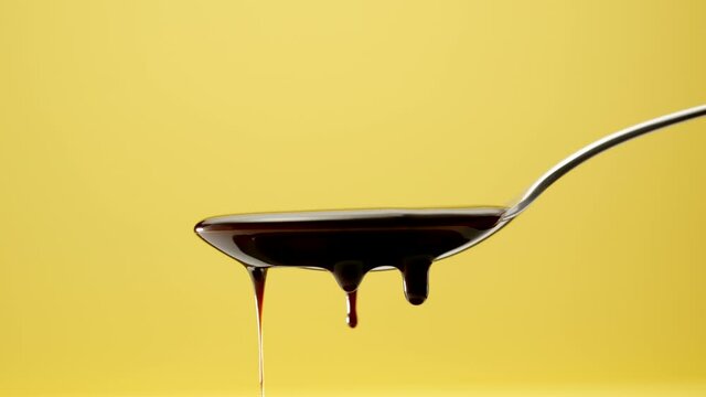 Drops of melted chocolate are dripping from a steel spoon on a yellow background, slow motion