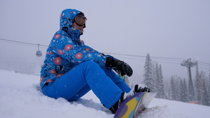 Fototapeta na wymiar Pretty young woman sitting on mountain peak with her snowboard and enjoying weather during snowfall. Young woman on ski slope