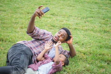Naklejka na ściany i meble Happy father with little son taking selfie picture with smartphone, Cheerful african american father and sons playing in park