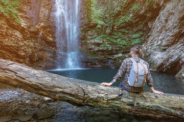 Fototapeta na wymiar Young tourist traveler with backpack resting on fallen tree trunk and enjoying view of waterfall.
