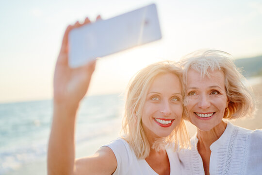 Outdoor portrait of smiling happy caucasian senior mother with her adult daughter taking selfie on smartphone on sea beach. - Powered by Adobe