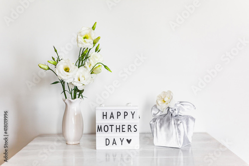 Gift box trendy wrapped in silk fabric in Furoshiki technique, light box with lettering Happy Mother's Day, white flowers Eustoma or Lisianthus in vase on wooden background. Zero Waste Life Concept