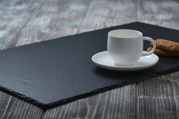 White cup for coffee on a black textured stone plate, cookies, wooden textured table, side view.