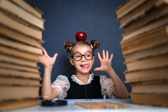 Happy smart girl in rounded glasses with red apple on her head sitting between two piles of books, have fun and look at camera smiling. - Powered by Adobe
