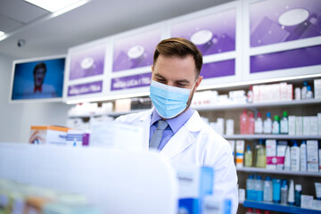 Pharmacist wearing face mask and white coat working in pharmacy store during corona virus pandemic arranging medicines on the shelf. Healthcare and medicine.