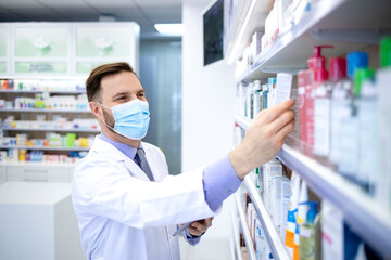 Pharmacist in protection mask holding tablet computer and checking medicines in pharmacy shop.