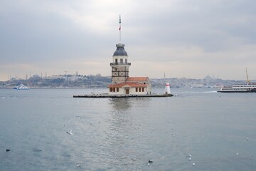 Fototapeta na wymiar The Maiden's Tower under gray cloudy sky, Bosphorus, Istanbul, Turkey during overcast weather with sunshine reflection in bosporus sea. Groups of seagulls flying on sea. İstanbul Turkey 01.03.2021