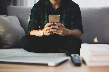 An Asian male freelance wear a plaid shirt sitting at work, posing comfortably on the sofa at home. He's checking and replying to emails and chatting with colleagues on his smartphone.