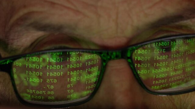 The Programmer with Wlasses in front of the Display. Reflection of the Monitor with a Numeric Code. The Light from the Computer Screen Reflects off the Glasses. Close-up of the Eyes.Pensive Focused