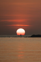 Sunset ocean view. Sun above the sea on orange sky. Fisherman with fishing rod on the rock.