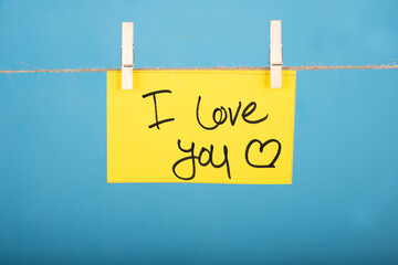 I Love You word written on a Yellow color sticky note hanging with a wire in front of Blue background.