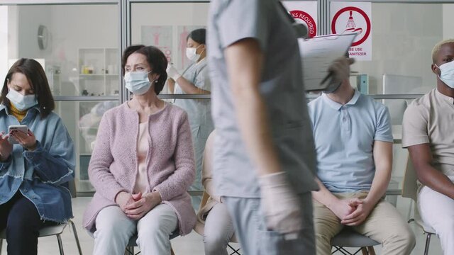 Slow-motion medium shot of multiethnic people sitting on chairs in queue at modern hospital before getting vaccinated against coronavirus