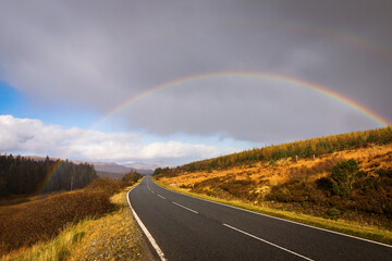 Autumn landscape in Highlands, Scotland, United Kingdom. Road with beautiful and colourful rainbow in background.