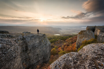 Fototapeta premium Silhouette of a woman on the top of a rock enjoys the view of sunset over an autumn forest