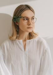 Young blonde woman in glasses, wearing a white blouse, standing at a white wall.