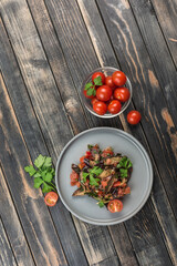 Warm salad with eggplant and tomatoes on a light flat plate and cherry tomatoes on a dark wooden background.