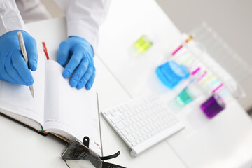 Fototapeta premium Employee of the chemical laboratory teacher chemist holds a silvery pen in his hand makes notes in the diary records test data from reactions examining test tubes with the substance arm in gloves