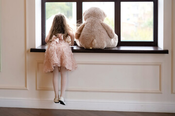 A beautiful little girl in a smart dress looks out the window, next to her favorite toy - a big teddy bear.