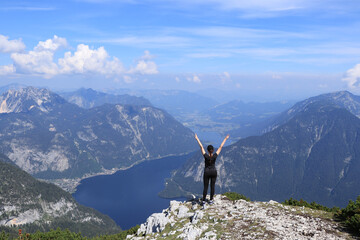 girl in black sportswear rejoices in defeating the Dachstein Krippenstein and enjoys the view of Hallstatter See and the adjacent village of Hallstatt with her hands over her head.