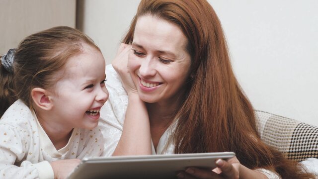 Happy mom and baby girl look at the tablet display and laugh while choosing purchases. Remote communication with relatives through the video camera of the gadget. Teaching a child online at home