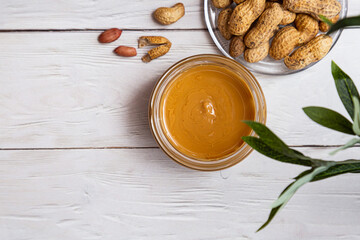 A jar of peanut butter with a bunch of peanuts on a white wooden table with an olive branch. Homemade peanut butter, natural, healthy food. The modern wellness and vegan concept. 